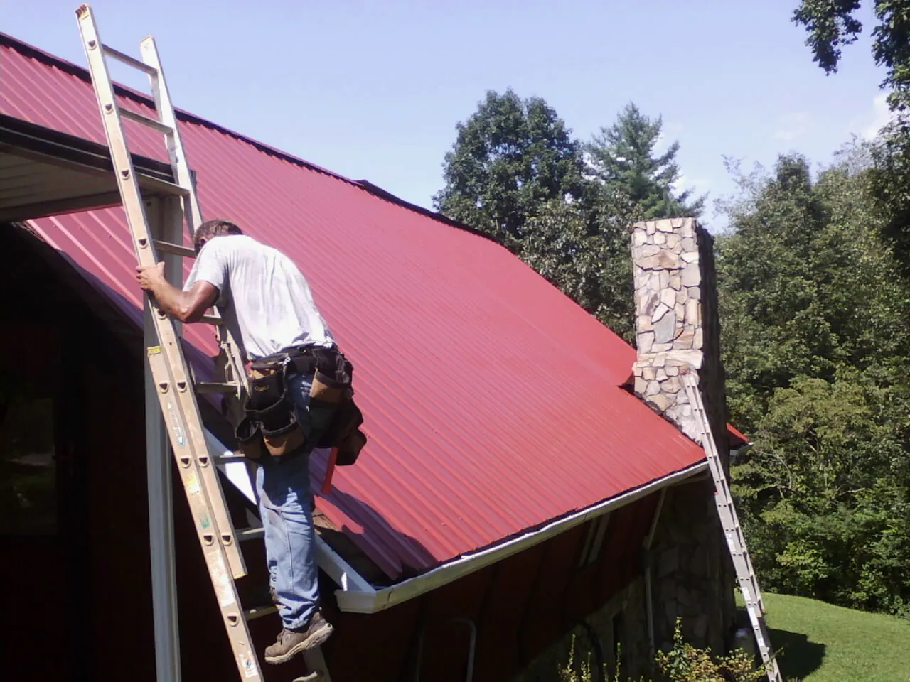 Roofer on a ladder beside a red metal roof and stone chimney — Soffit & Fascia Repair by Roofing Brothers in Mifflin